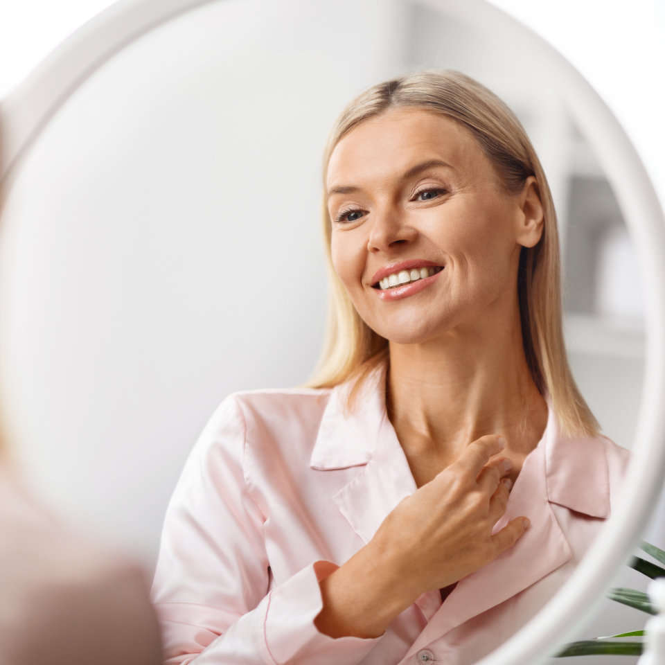 A woman with long hair and a pink shirt smiles while looking at herself in a round mirror.