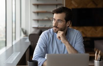 Thoughtful businessman in glasses looking at window from workplace with laptop, thinking over challenges, work issues, problem solving, making decision, feeling doubts, pondering on future vision